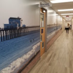 Custom 60-foot hallway wall mural installation depicting surfers in ocean near long pier with restaurant on the end