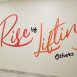 "You Rise by Lifting Others" large cut vinyl wall letters in cursive.