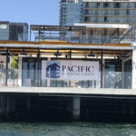 up close view from San Diego Bay of the new Portside Pier being built on pylons over the bay with large banner for Pacific Building Group